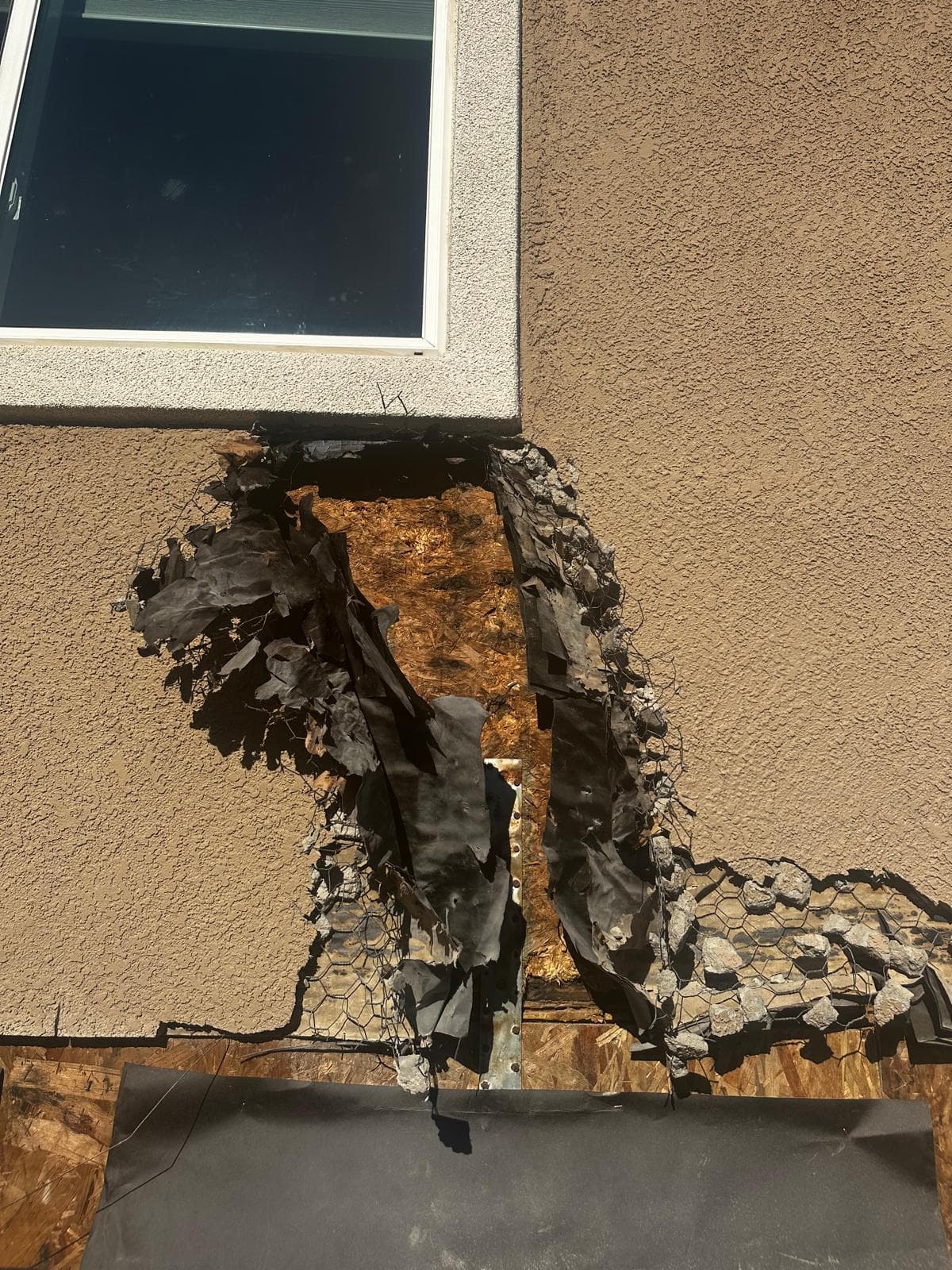 A large opening below a window in a stucco wall exposing torn black building paper, chicken-wire lath, and bare sheathing