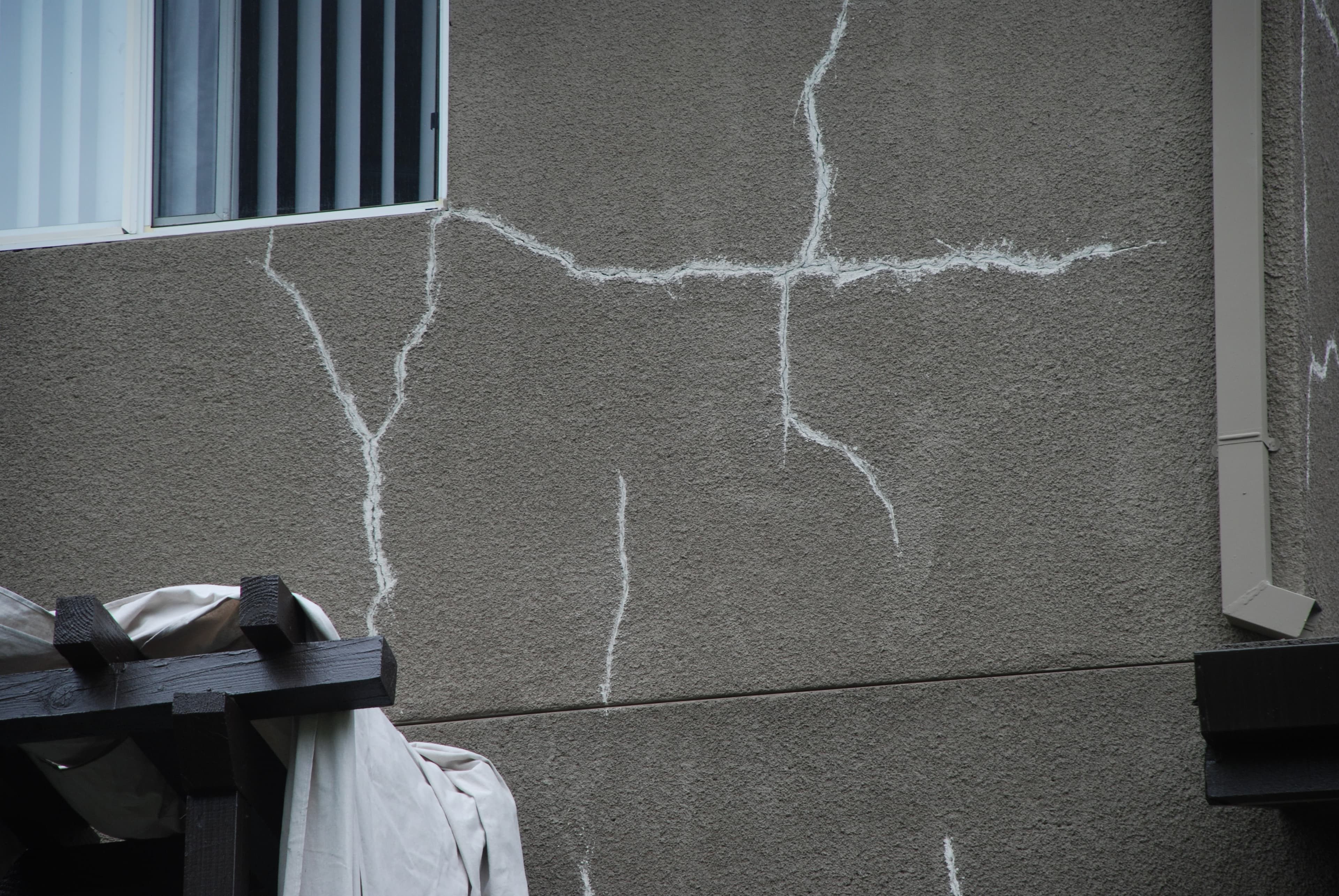 Branching stucco cracks spreading from the bottom corner of a window, running through the lighter lines of earlier patches
