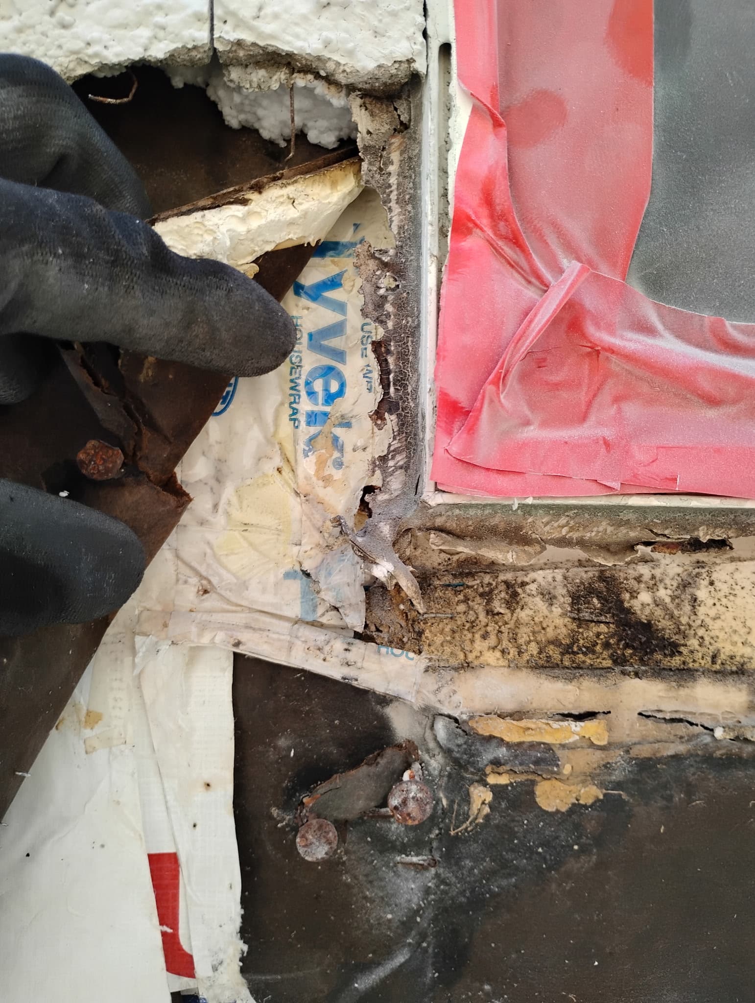Stucco removed at a window corner revealing rusted flashing, deteriorated building paper, and dark water damage behind the wall