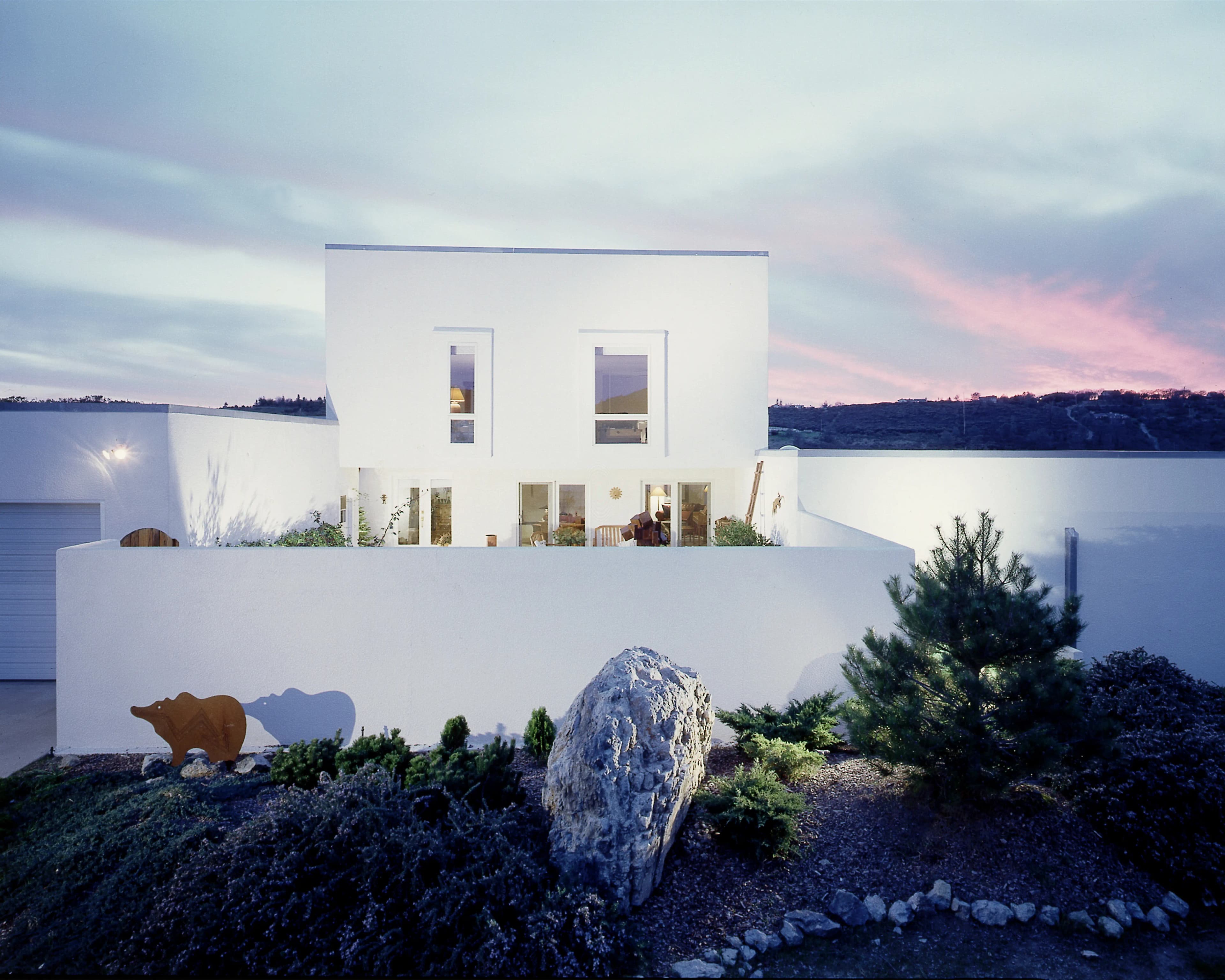 Contemporary white stucco home at dusk with warm interior lighting and a rock garden