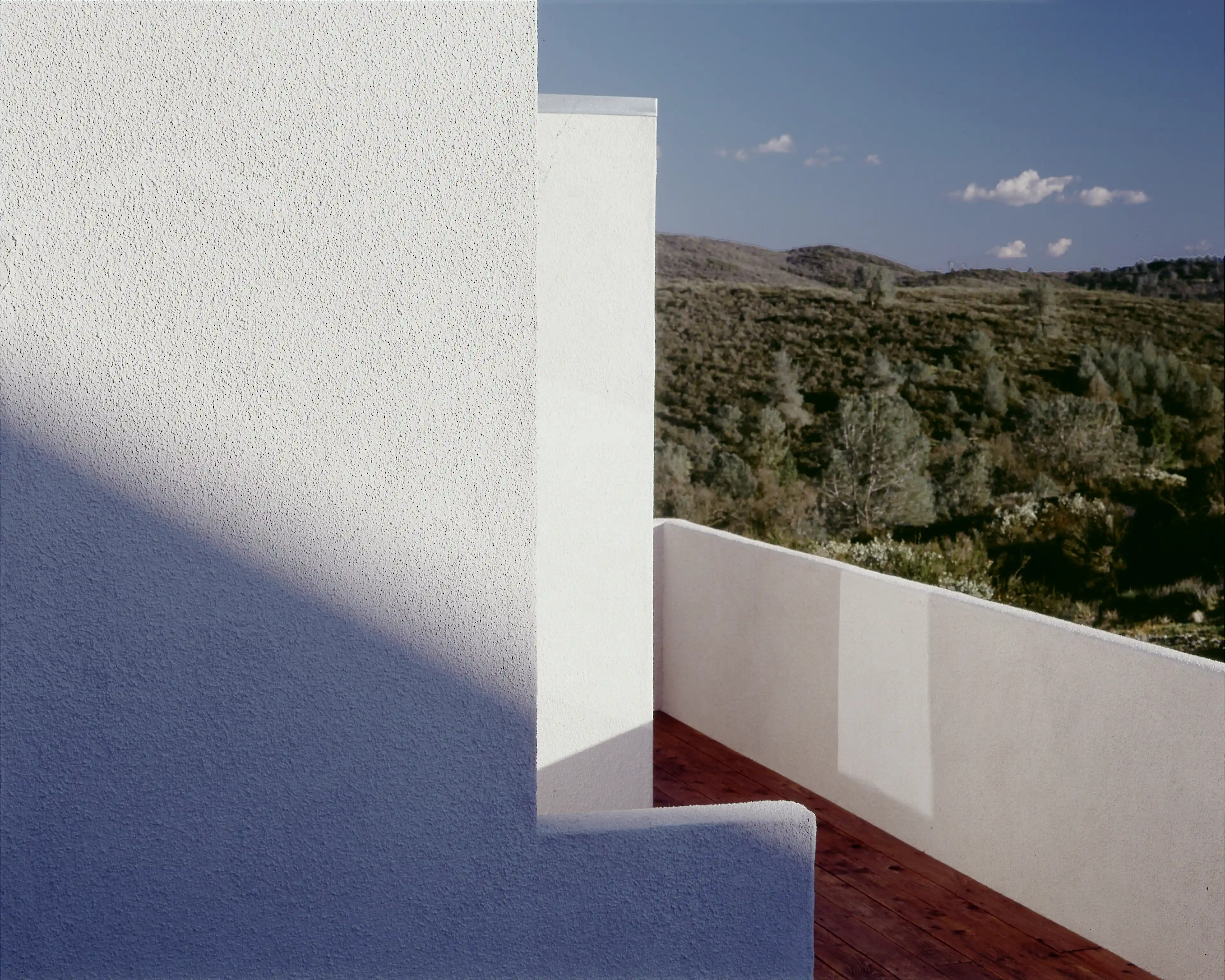 Sunlit white stucco wall and parapet meeting a redwood deck, with hillside vegetation beyond