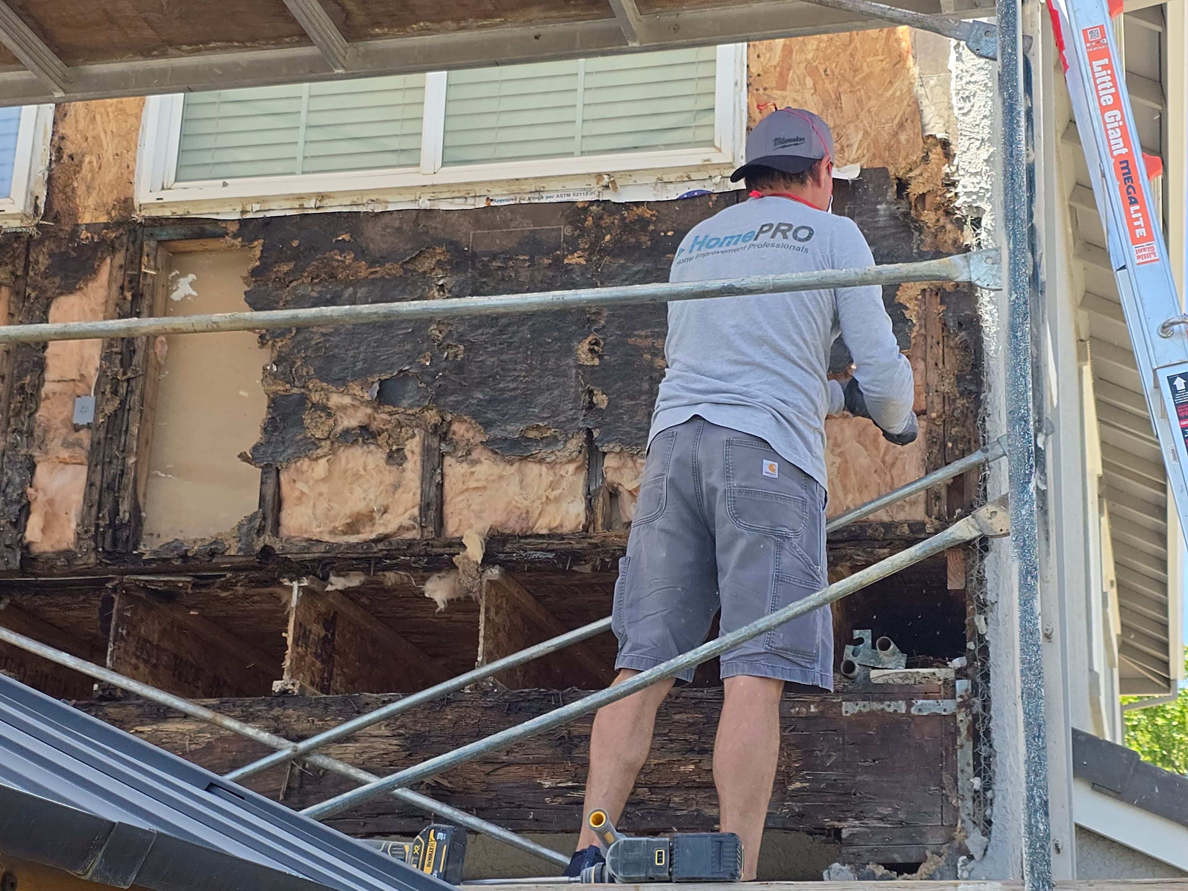 A contractor on scaffolding works on an exterior wall where siding has been removed, exposing decayed wood sheathing and framing around a window.